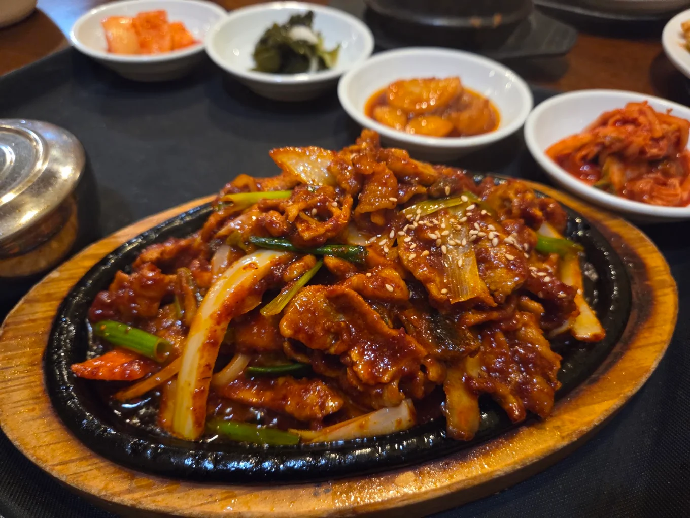 Spicy pork jeyuk sizzling on a cast-iron plate with sesame seeds, onion and scallion, surrounded by banchan bowls