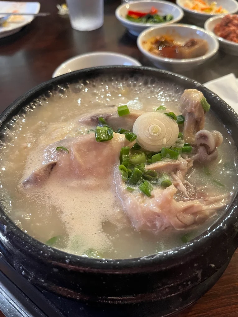 Samgyetang ginseng chicken soup bubbling in a hot stone pot, topped with scallion, banchan behind