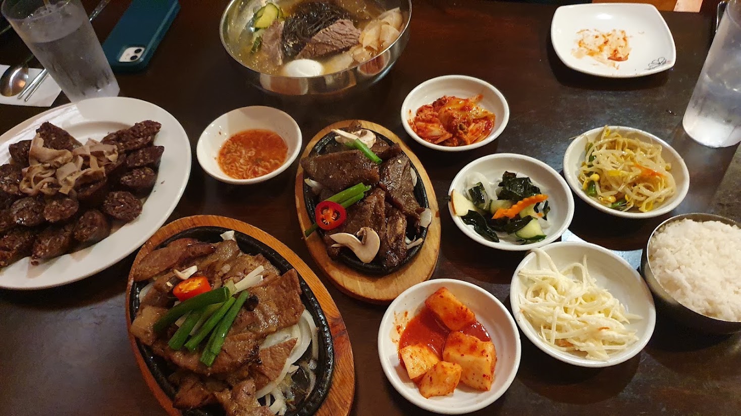 A full Korean table spread with sizzling grilled meats, sliced sundae, beef soup, rice and a ring of banchan dishes